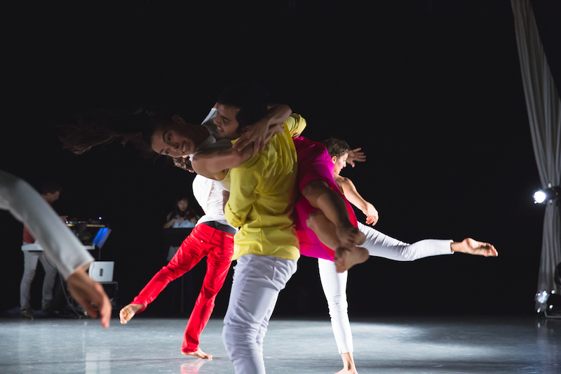 A dancer picks up another whose legs pike up around his back. Dancers whose legs kick up in the air are in the background.